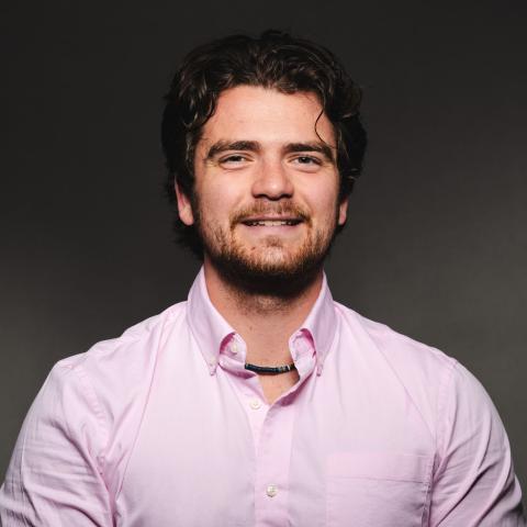 Aidan McKee wearing a pink button down shirt smiling for his headshot in front of a black backdrop.